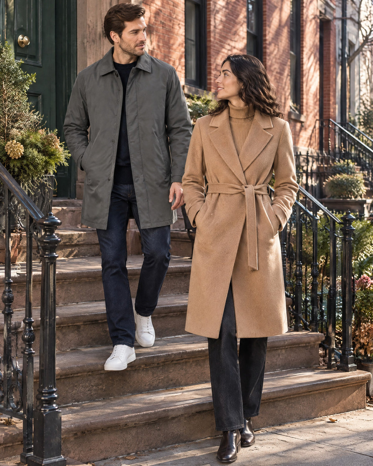 On a brownstone stoop with iron railings, a man in a gray car coat over a navy knit and dark jeans beside a woman in a camel belted wrap coat over a camel turtleneck
