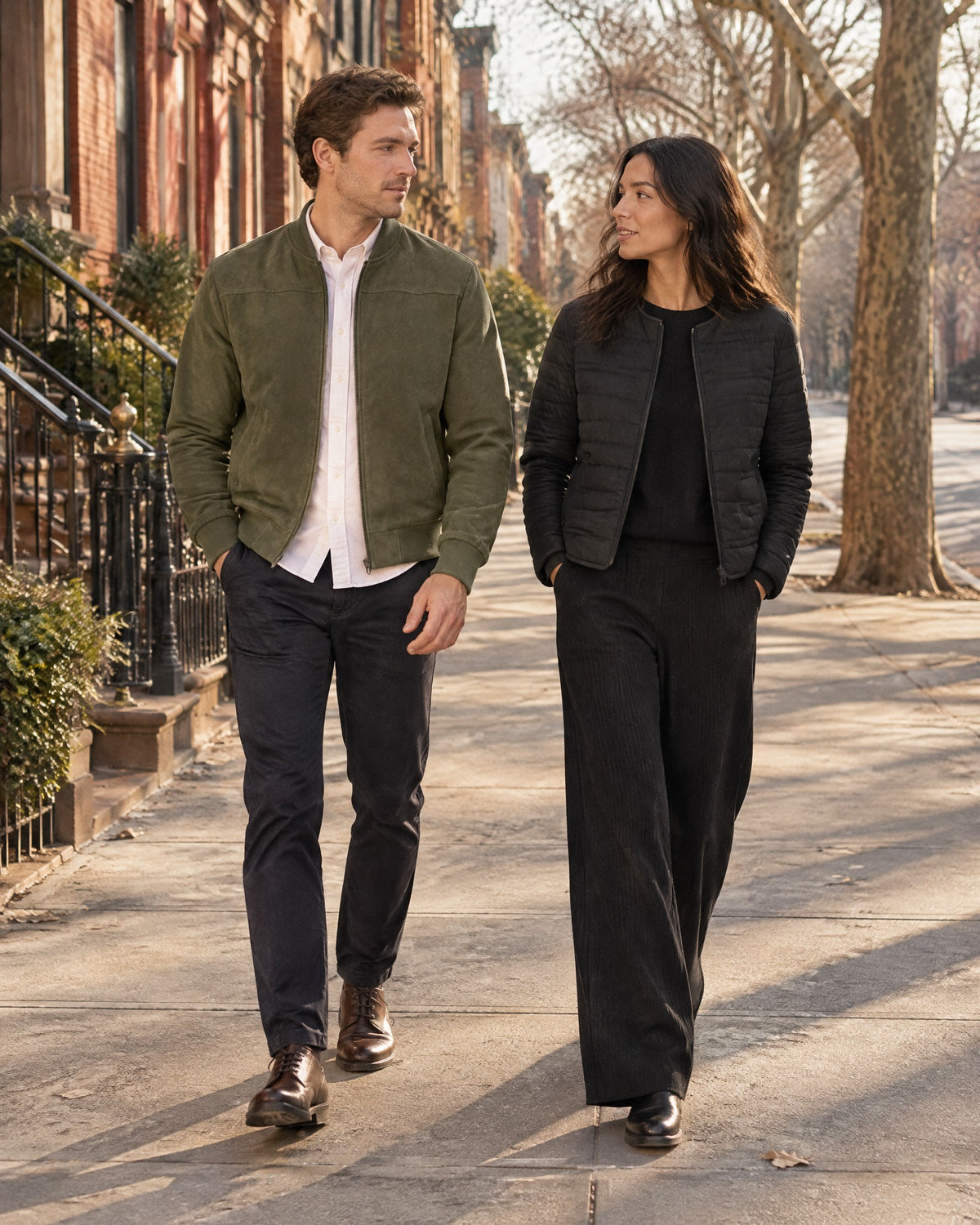 Two friends walk a tree-lined brownstone street in winter sun, he in an olive faux-suede bomber over a white oxford shirt, she in a black quilted jacket and black ribbed wide-leg trousers