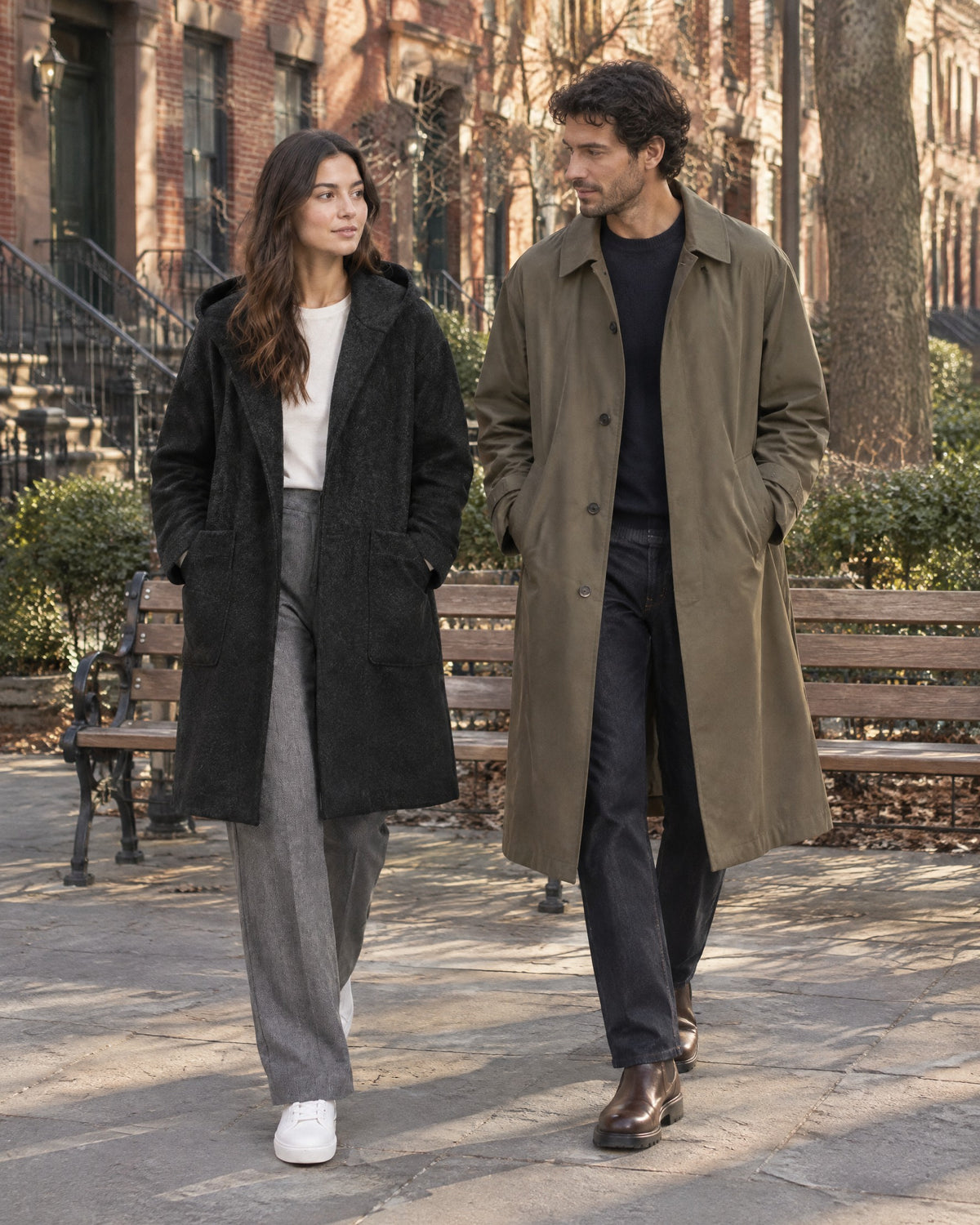 In a small square by a wooden bench, a woman in a black hooded coat over gray trousers and a man in a long khaki trench coat over a navy knit